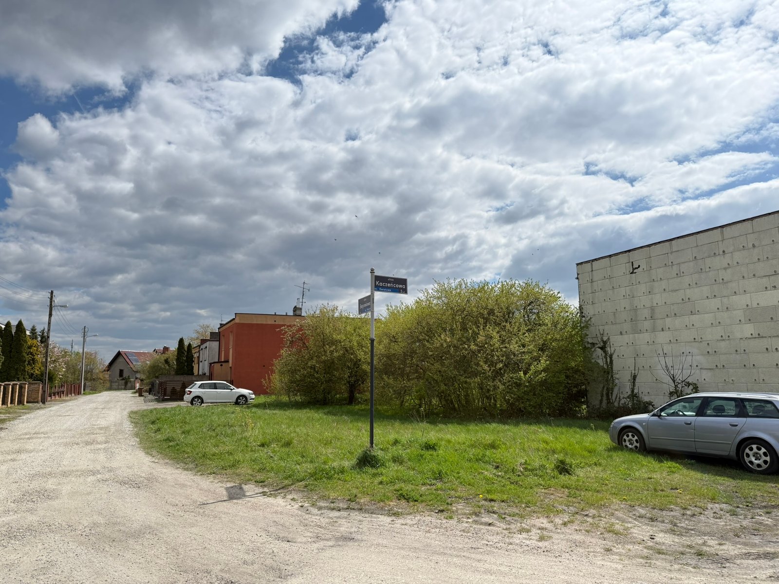 Widok na teren działki od ul. Kaczeńcowej, niezabudowana, porośnięta niską roślinnością i krzewami.