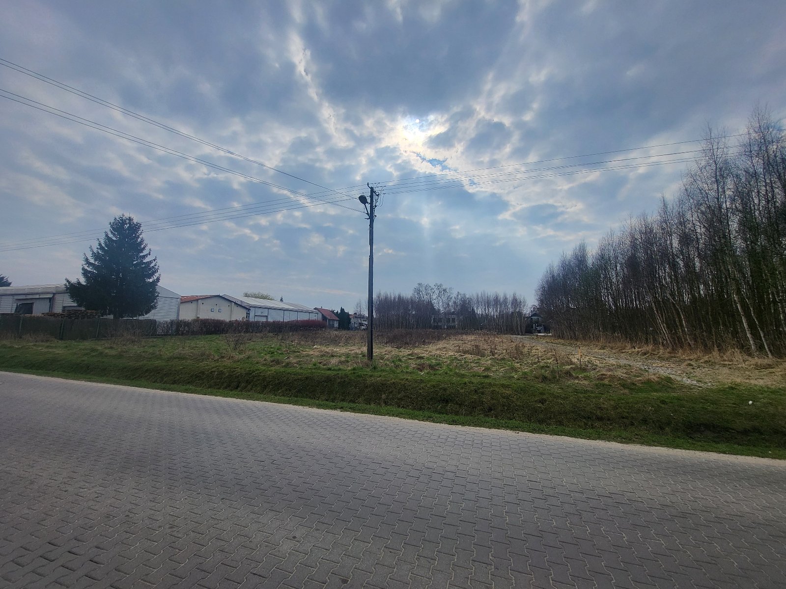 Zdjęcie przedstawia działkę budowlaną, położoną w Poznaniu przy ul. Sieweczki, działka ta jest delikatnie zarośnięta wysoką trawą, widoczny jest również słup na granicy działki oraz rów melioracyjny od strony ulicy Zielińskiej w miejscowości Gruszczyn oraz ulica Sieweczki
